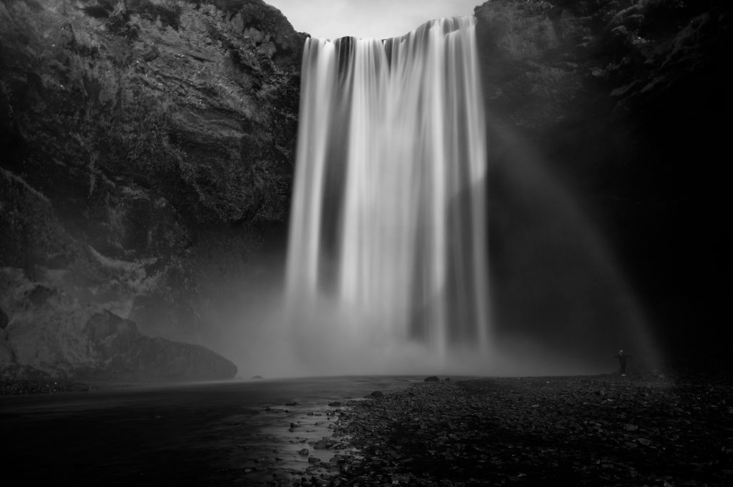 Schwarz-weiß Aufnahme eines Wasserfalls mit Regenbogen und Nebel im Vordergrund.