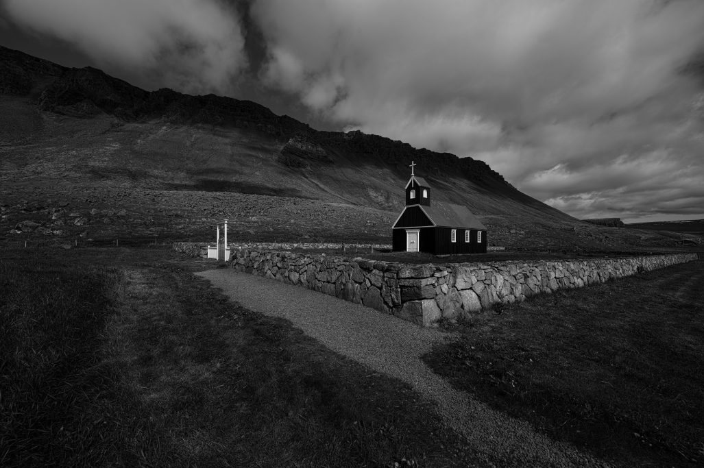 Schwarzes Kirchenggebäude vor einer hügeligen Landschaft unter bewölktem Himmel.