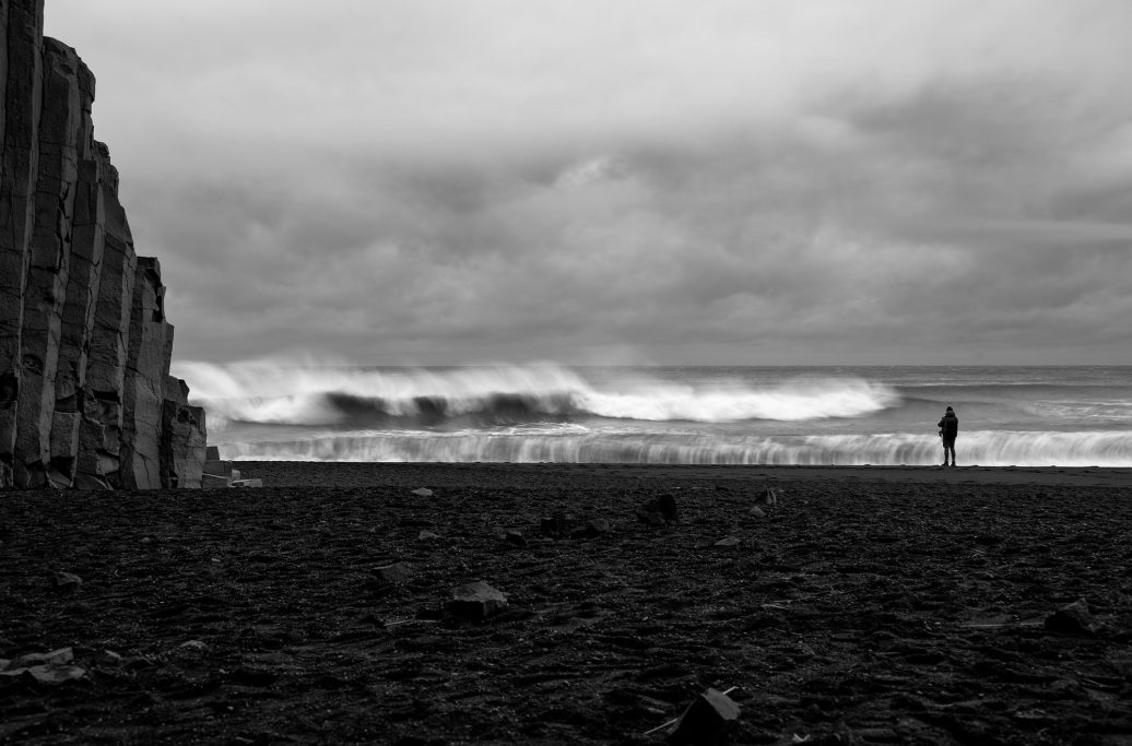 Düsterer Strand mit Wellen und einer einsamen Figur am Horizont.